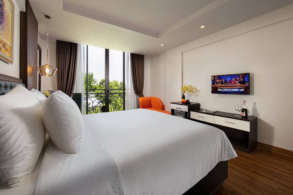 A hotel room with a large bed, an orange chair, a desk with a TV, and a window with curtains.
