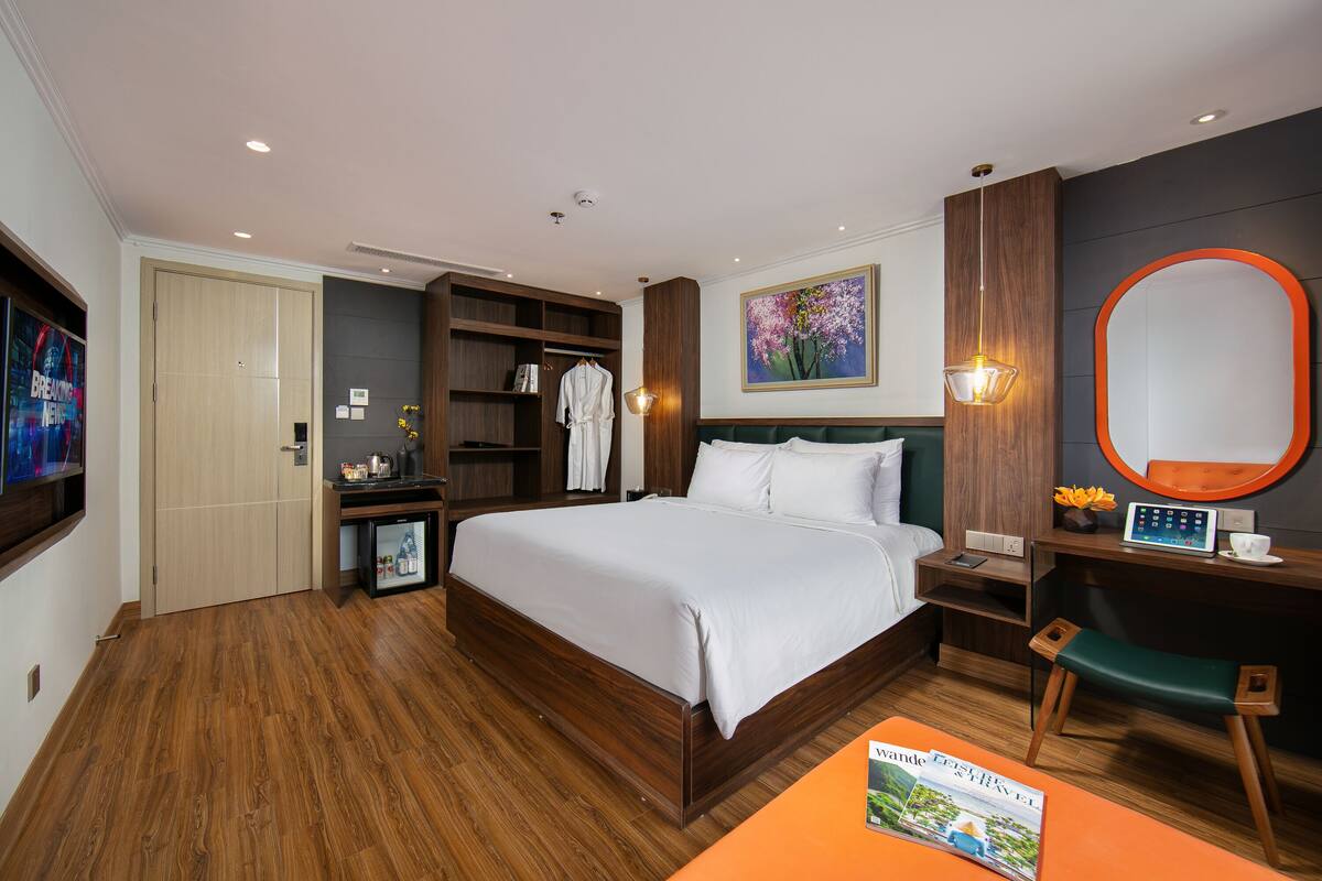 A hotel room with a large bed, a desk with a chair, a wardrobe, a small table with a vase, and a painting on the wall.