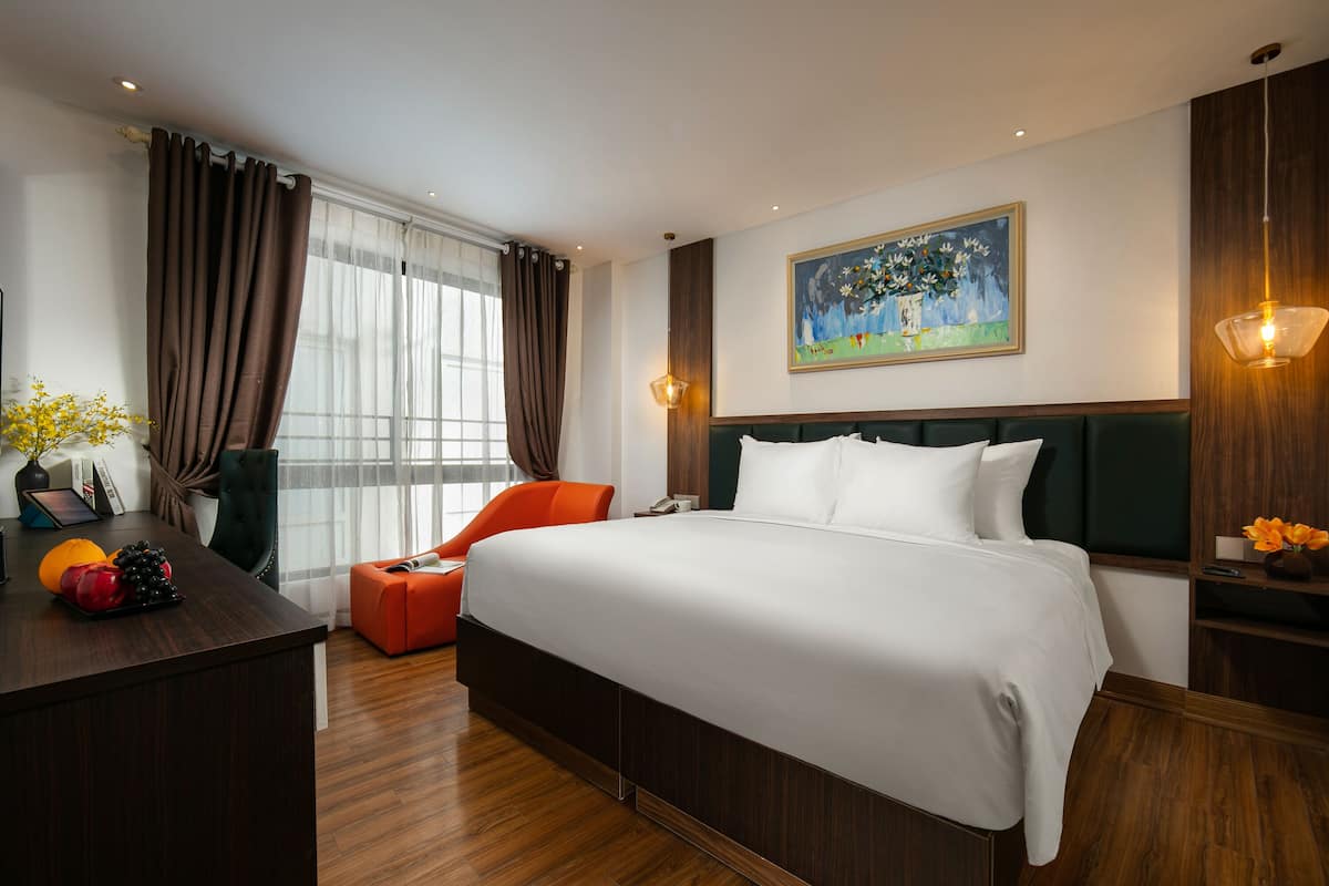 A modern hotel room with a large bed, a desk with fruit, a chair, and a painting on the wall.