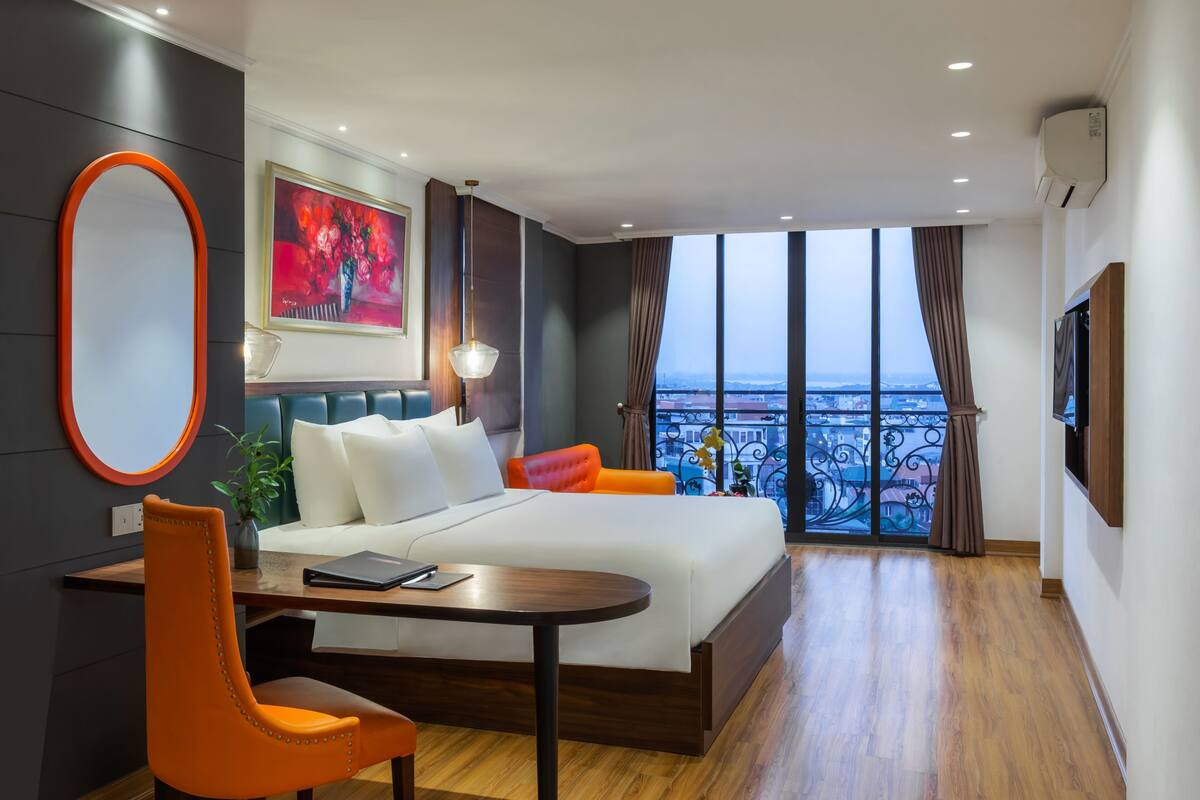 A hotel room with a large bed, a desk, an orange chair, a painting, and a view of the city through large windows.