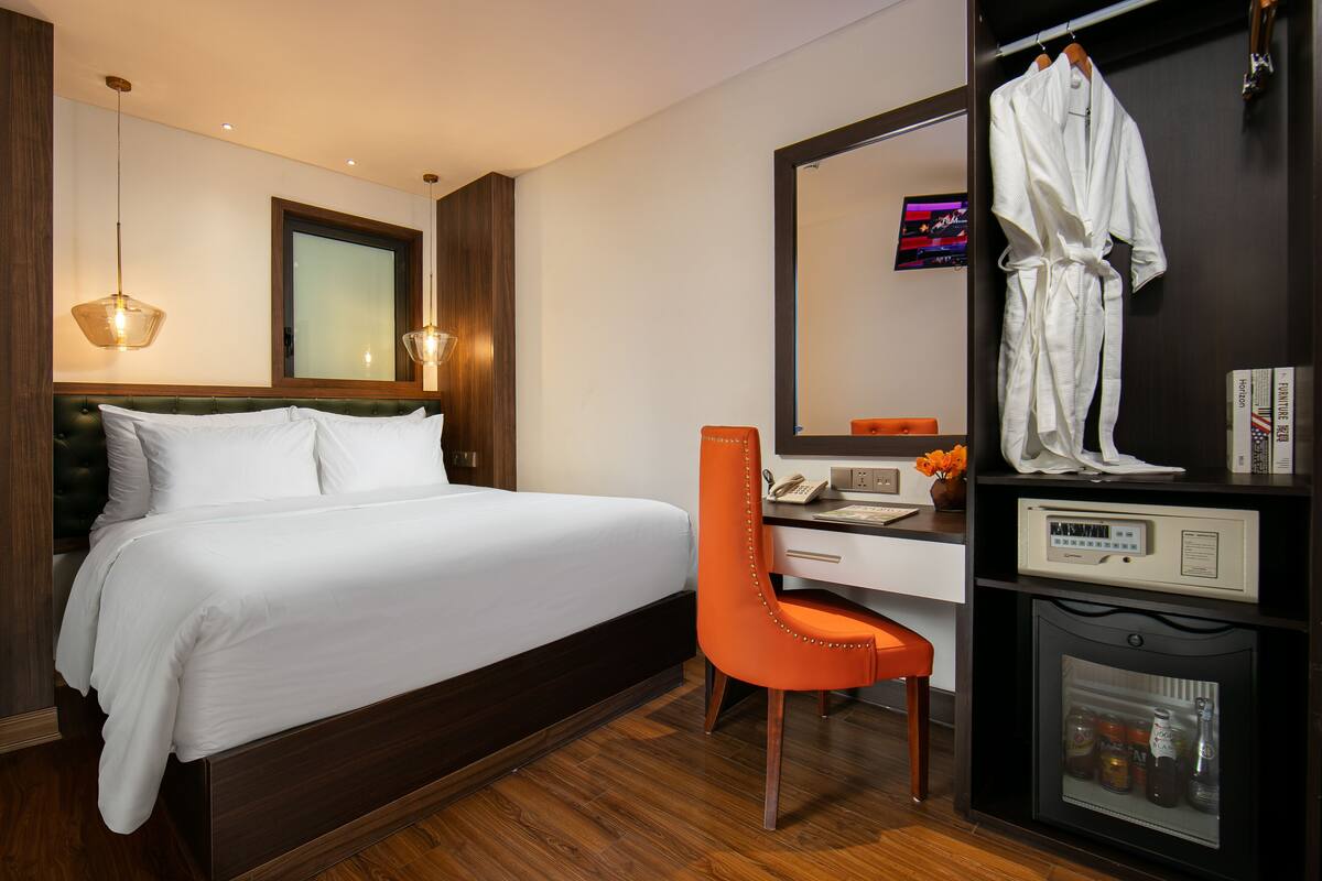 A hotel room with a bed, an orange chair, a desk with a telephone, a mirror, and a minibar.