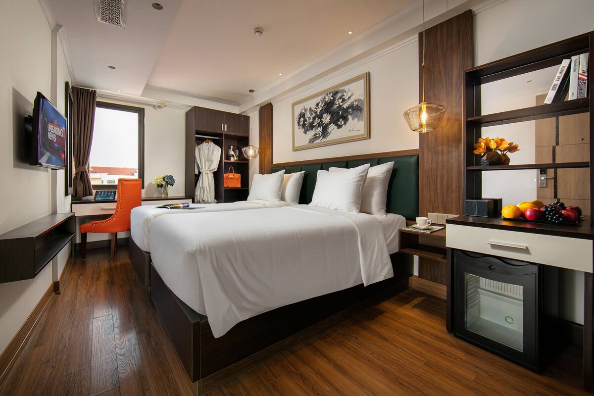 A hotel room with a large bed, a desk, a chair, a TV, a bookshelf, and a small refrigerator.