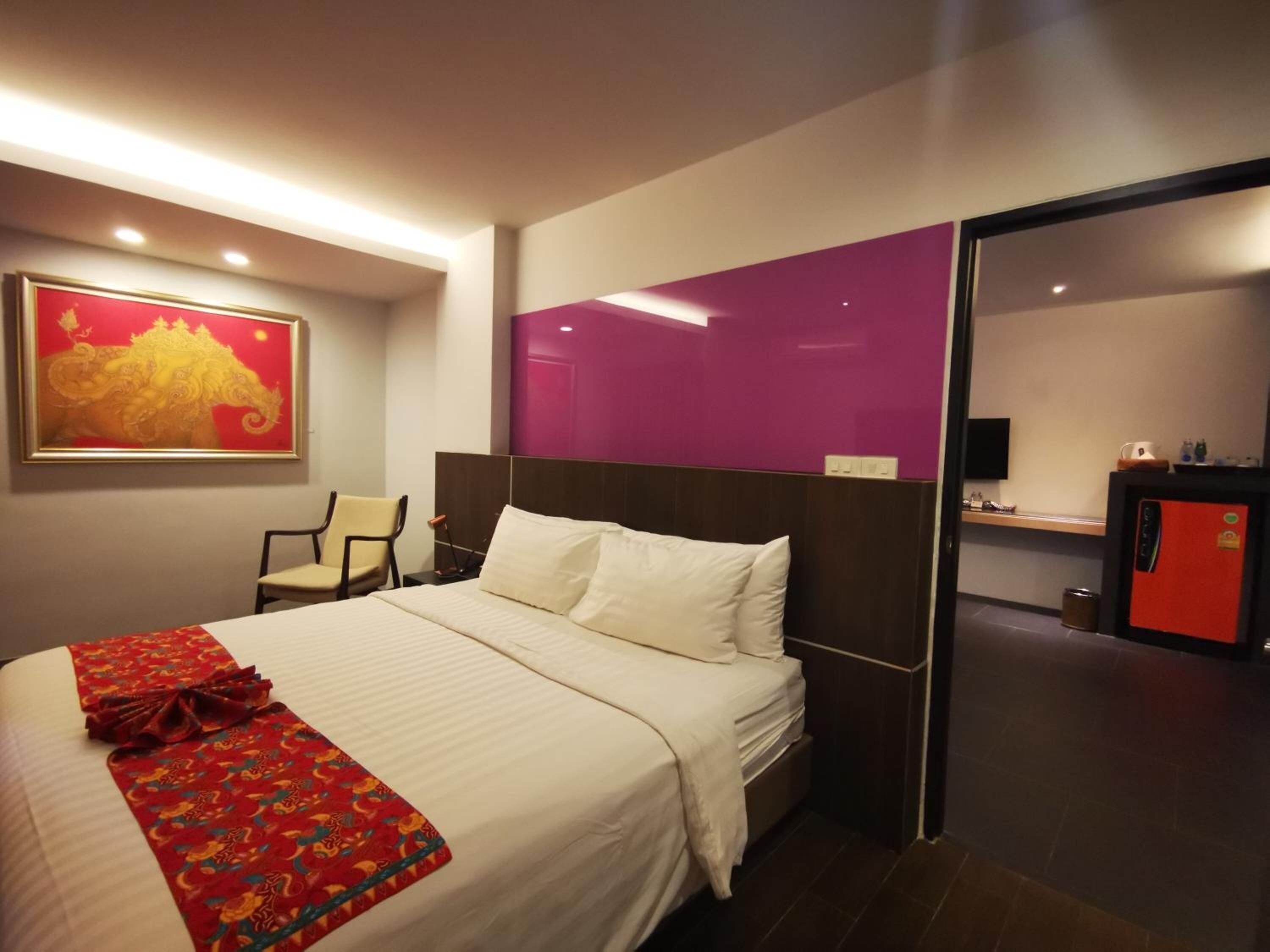 A hotel room with a bed, a chair, a desk, and a red refrigerator.