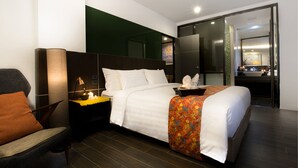 Habitación de hotel moderna con una cama grande, una silla, una mesita de noche y un baño visible a través de una puerta abierta.