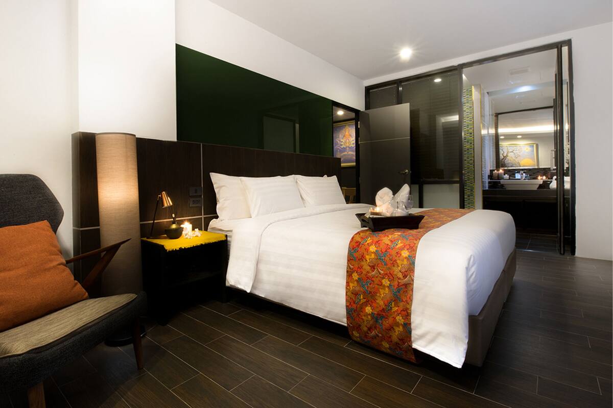 Habitación de hotel moderna con una cama grande, una silla, una mesita de noche y un baño visible a través de una puerta abierta.