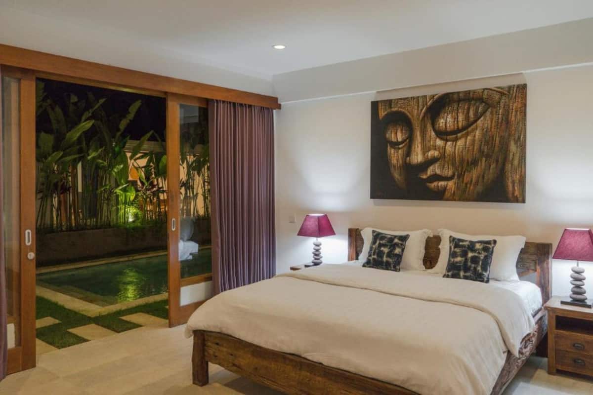 Quarto com uma cama grande, cabeceira de madeira, um quadro na parede e uma porta de vidro corrediça que dá para uma área de piscina.