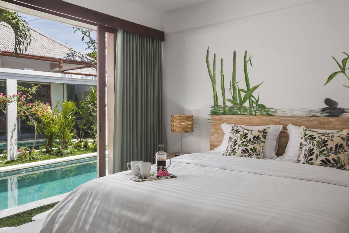 Quarto de hotel com uma cama, mesa de cabeceira com cafeteira, chávenas e um tabuleiro, uma janela ampla com vista para uma piscina e vegetação, e uma parede decorada com motivos de bambu.