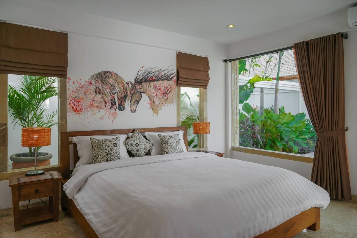 Quarto com uma cama grande, um mural na parede, uma janela com cortinas e uma mesa de cabeceira com abajur.