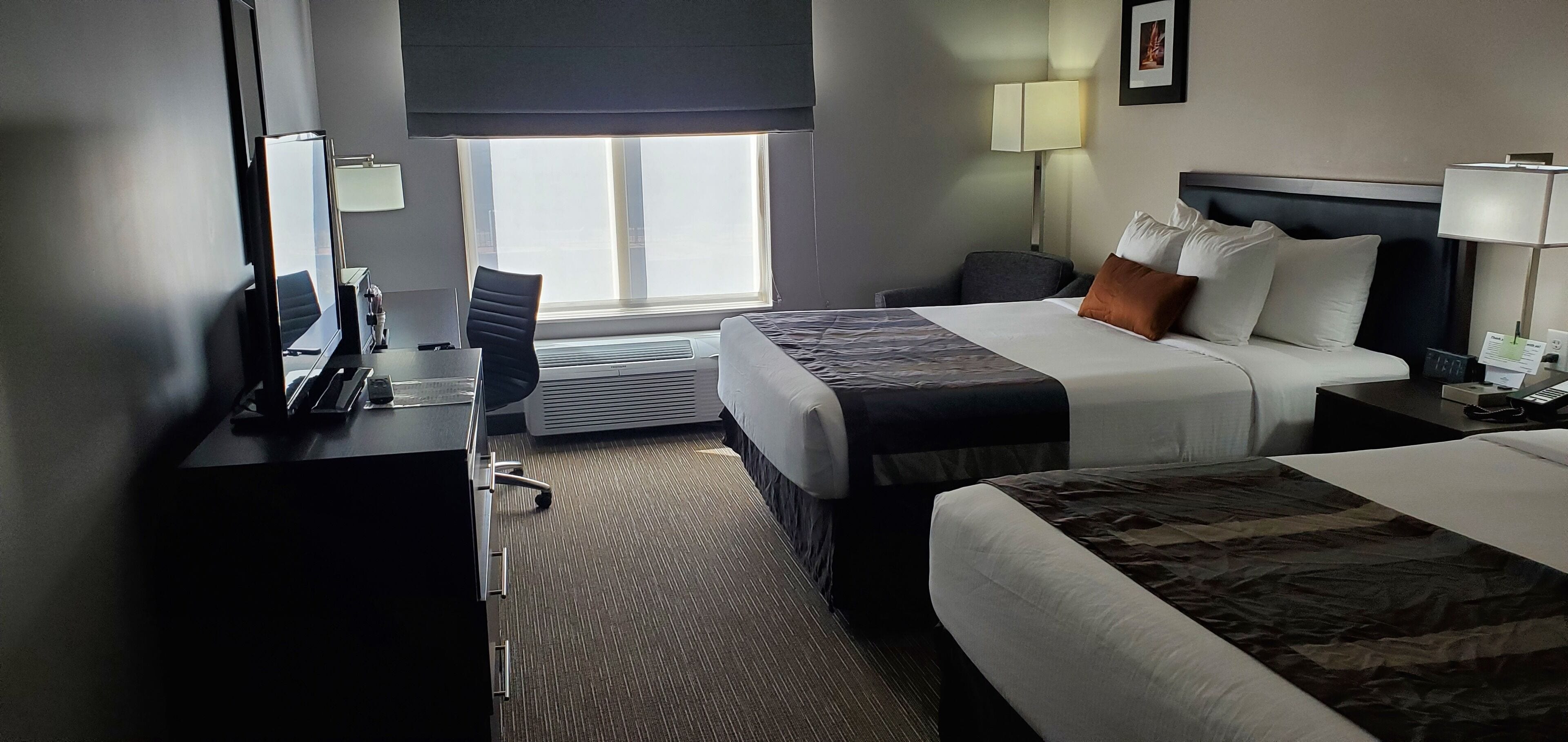 Quarto de hotel com duas camas, uma escrivaninha com cadeira, uma luminária e uma janela com vista.