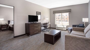 A hotel room with a flat-screen TV, a desk, a chair, a sofa, and a large window with a view of buildings.