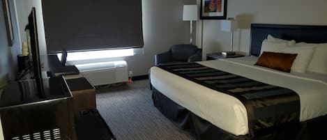 Ein Hotelzimmer mit einem Bett, einem Schreibtisch mit Computer, einem Sessel, einem Nachttisch und einem Fenster mit Jalousie.
