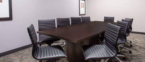 Un salón de conferencias con una gran mesa rectangular rodeada por ocho sillas de cuero negro con estructura metálica. El salón tiene piso alfombrado con un diseño geométrico y paredes de color gris claro.