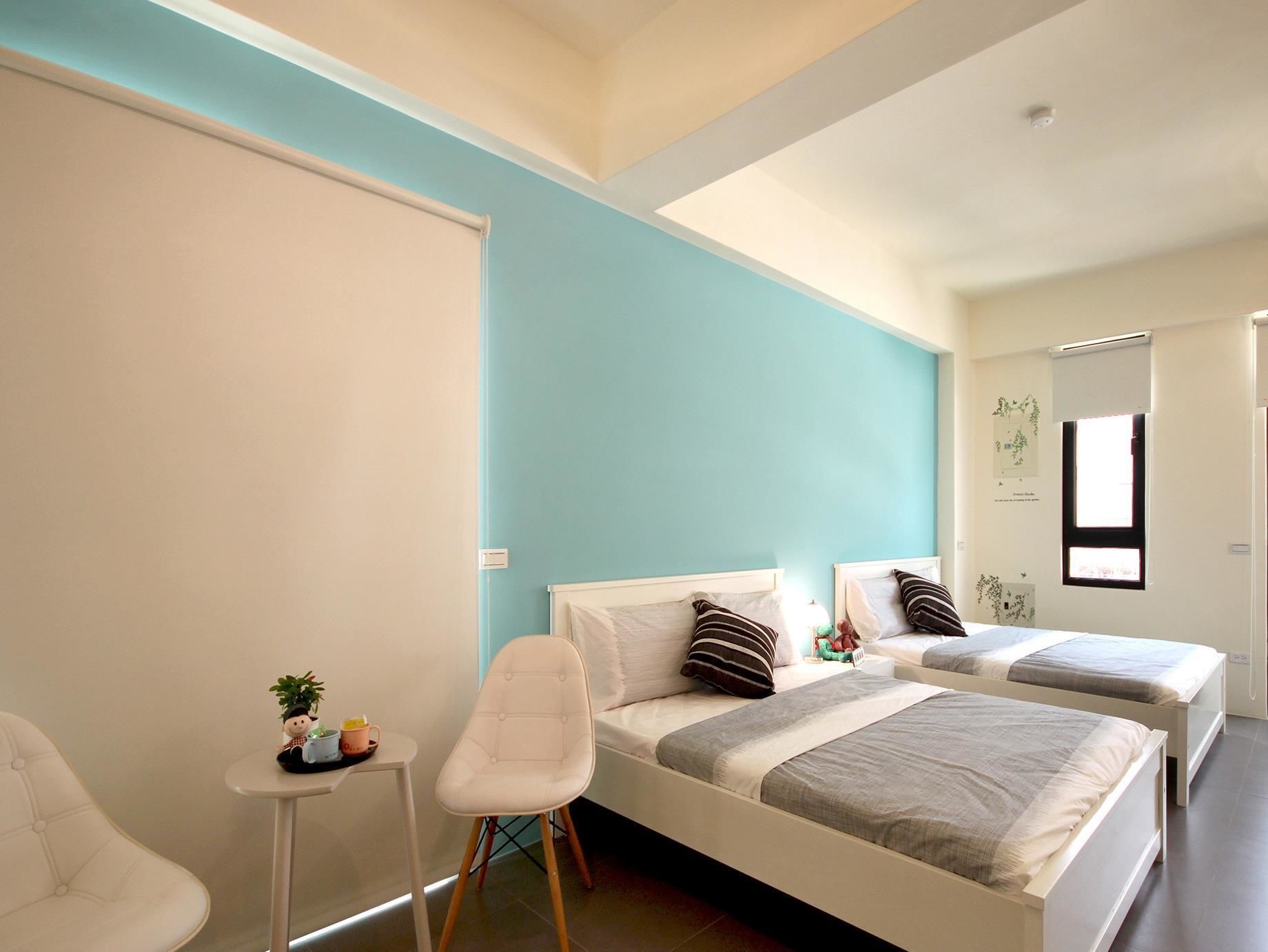 A modern hotel room with two beds, a small table with a chair, and a window with blinds.