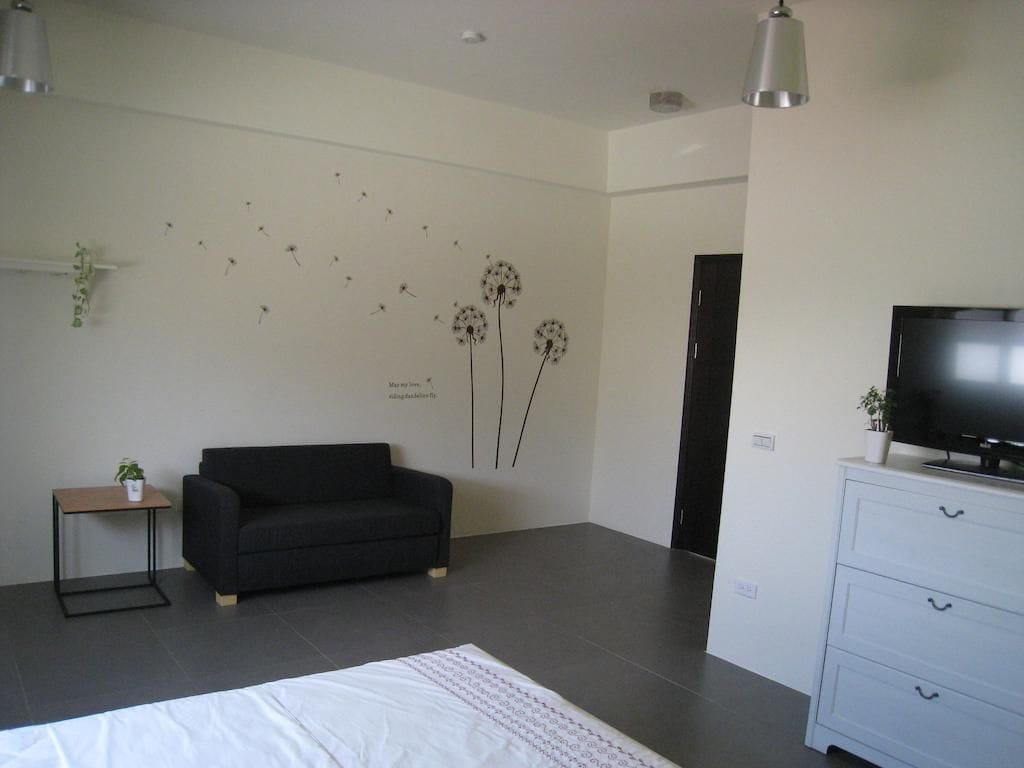 Un dormitorio moderno con un sofá negro, una cama con colcha blanca, un televisor sobre un aparador blanco y arte mural de diente de león.