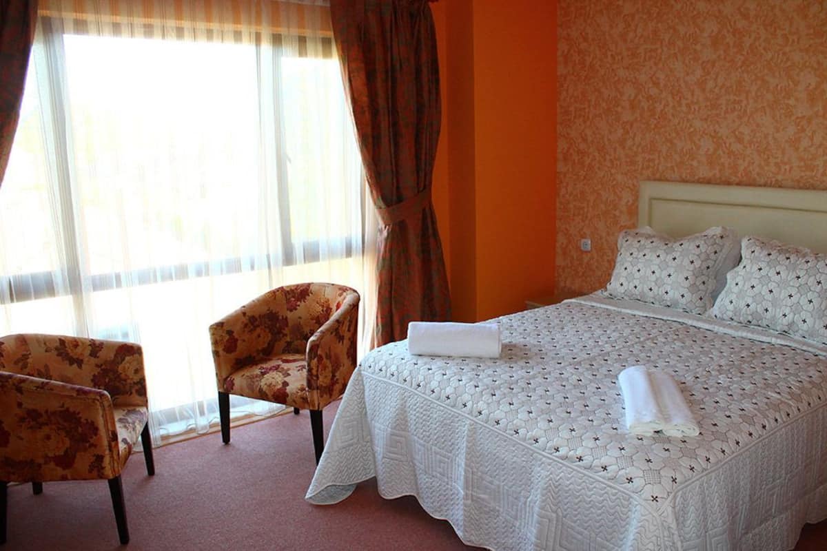 A hotel room with a large bed, two armchairs, a window with curtains, and an orange wall.