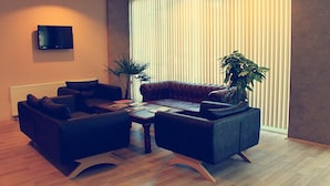 A modern living room with a leather sofa, two armchairs, a coffee table, and a TV mounted on the wall. There are two potted plants and a large window with blinds. - The Hotel Sapanca (Sapanca)