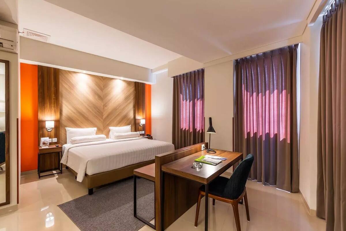 A hotel room with a bed, bedside table, lamp, desk, chair, and curtains.