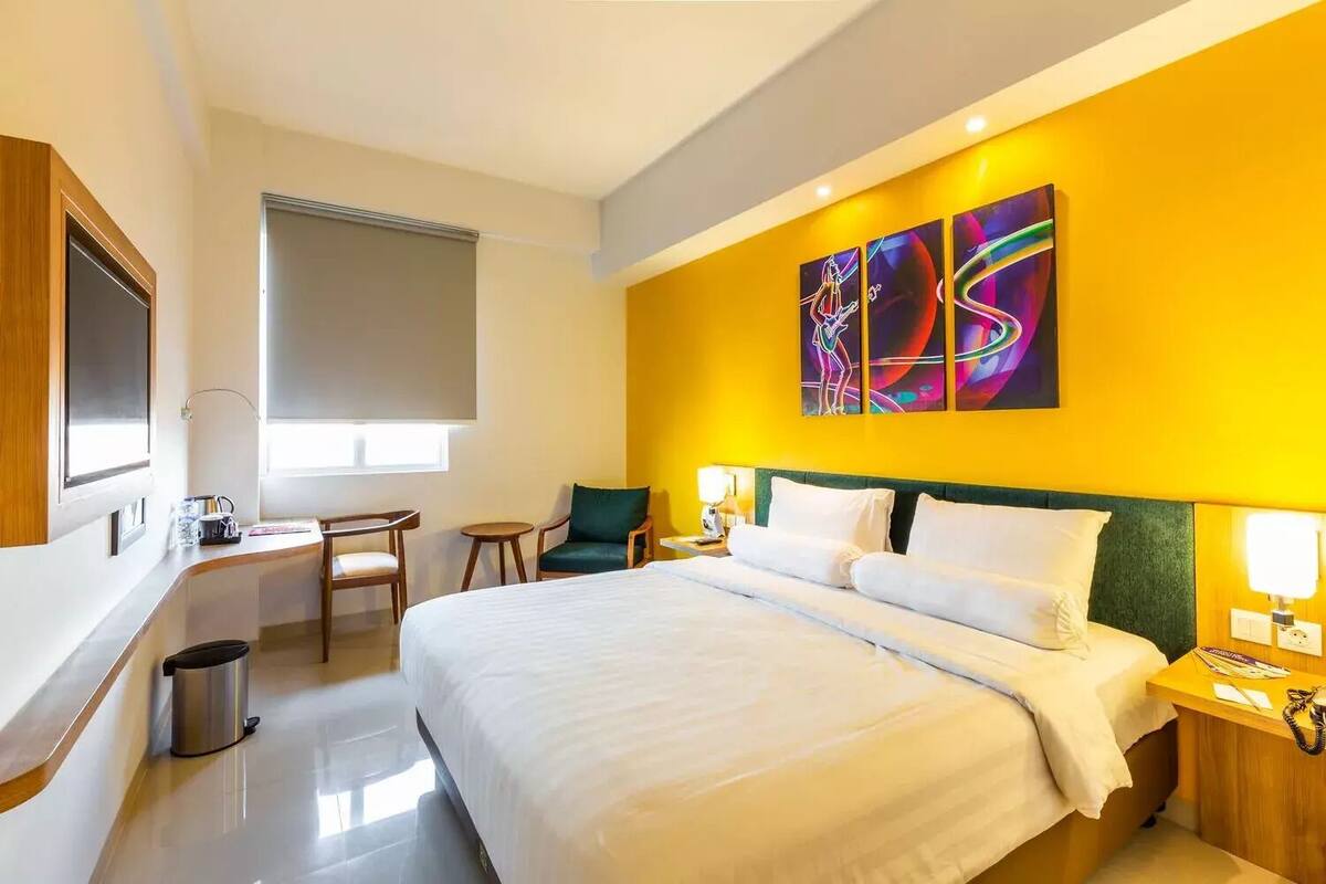 A hotel room with a large bed, a desk with a chair, a trash bin, and a window with blinds.