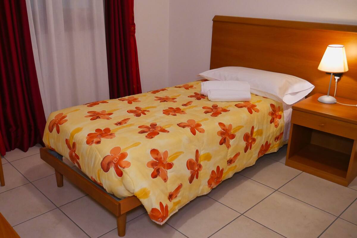 A hotel room with a bed, a nightstand with a lamp, a chair, and curtains.