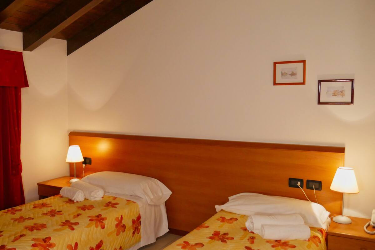 A hotel room with two beds, a wooden headboard, two bedside lamps, a red curtain, and two framed pictures on the wall.