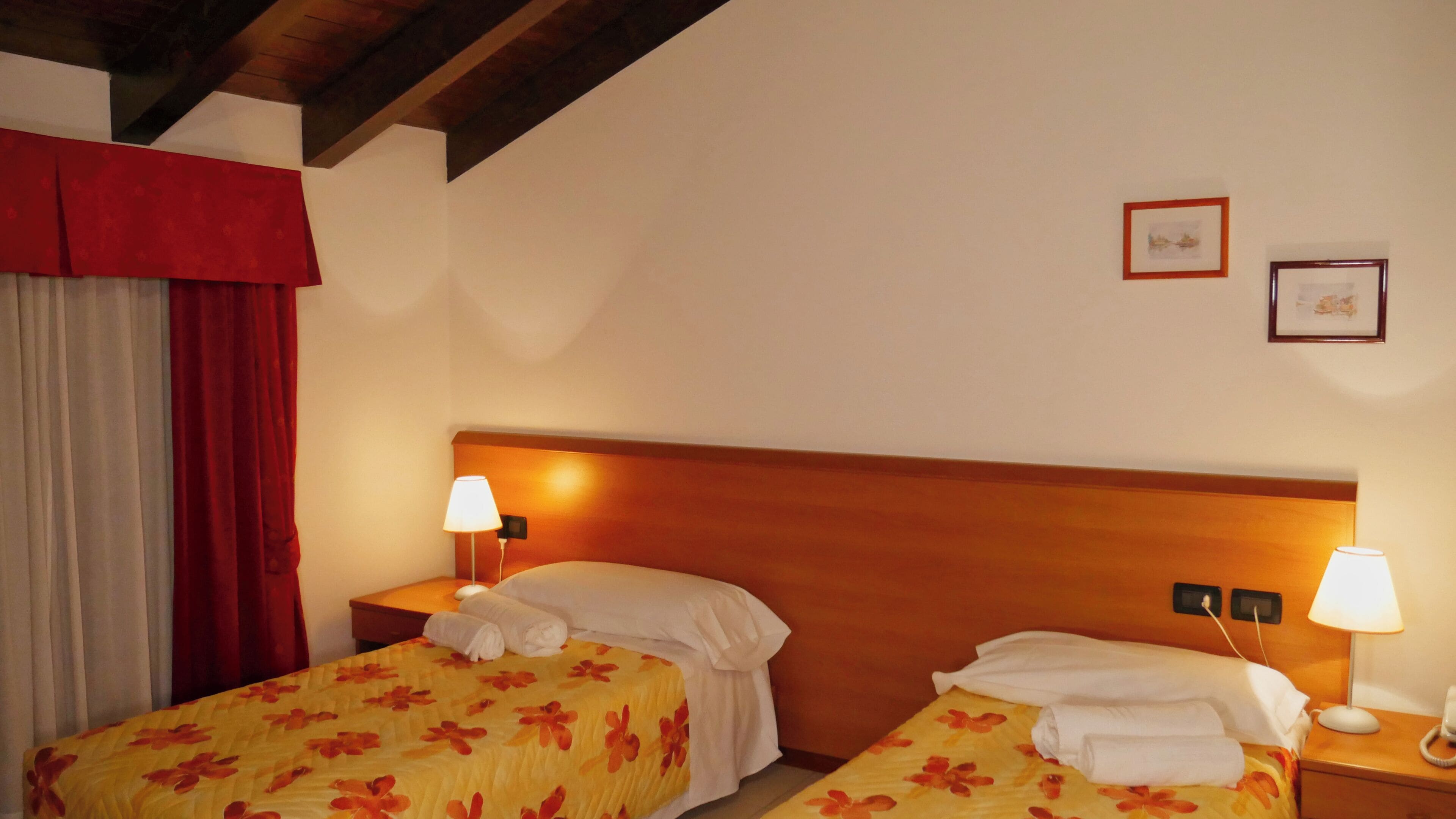 A hotel room with two beds, a wooden headboard, two bedside lamps, a red curtain, and two framed pictures on the wall.