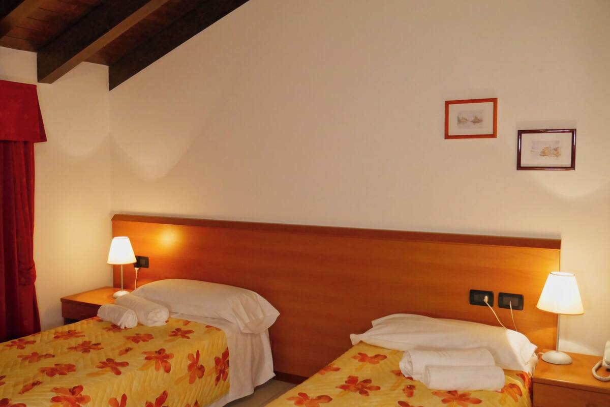 A hotel room with two beds, a wooden headboard, two bedside lamps, a red curtain, and two framed pictures on the wall.