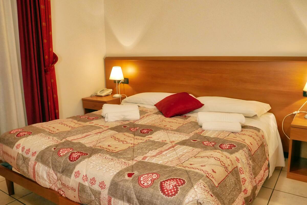 A neatly made bed with a patterned bedspread, two bedside lamps, a wooden headboard, and a red pillow.