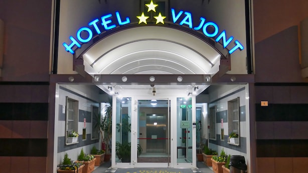 The entrance of Hotel Vaillant with a checkered floor mat, glass doors, and potted plants. - Hotel Vajont (Vajont)
