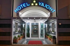 The entrance of Hotel Vaillant with a checkered floor mat, glass doors, and potted plants. - Hotel Vajont (Vajont)
