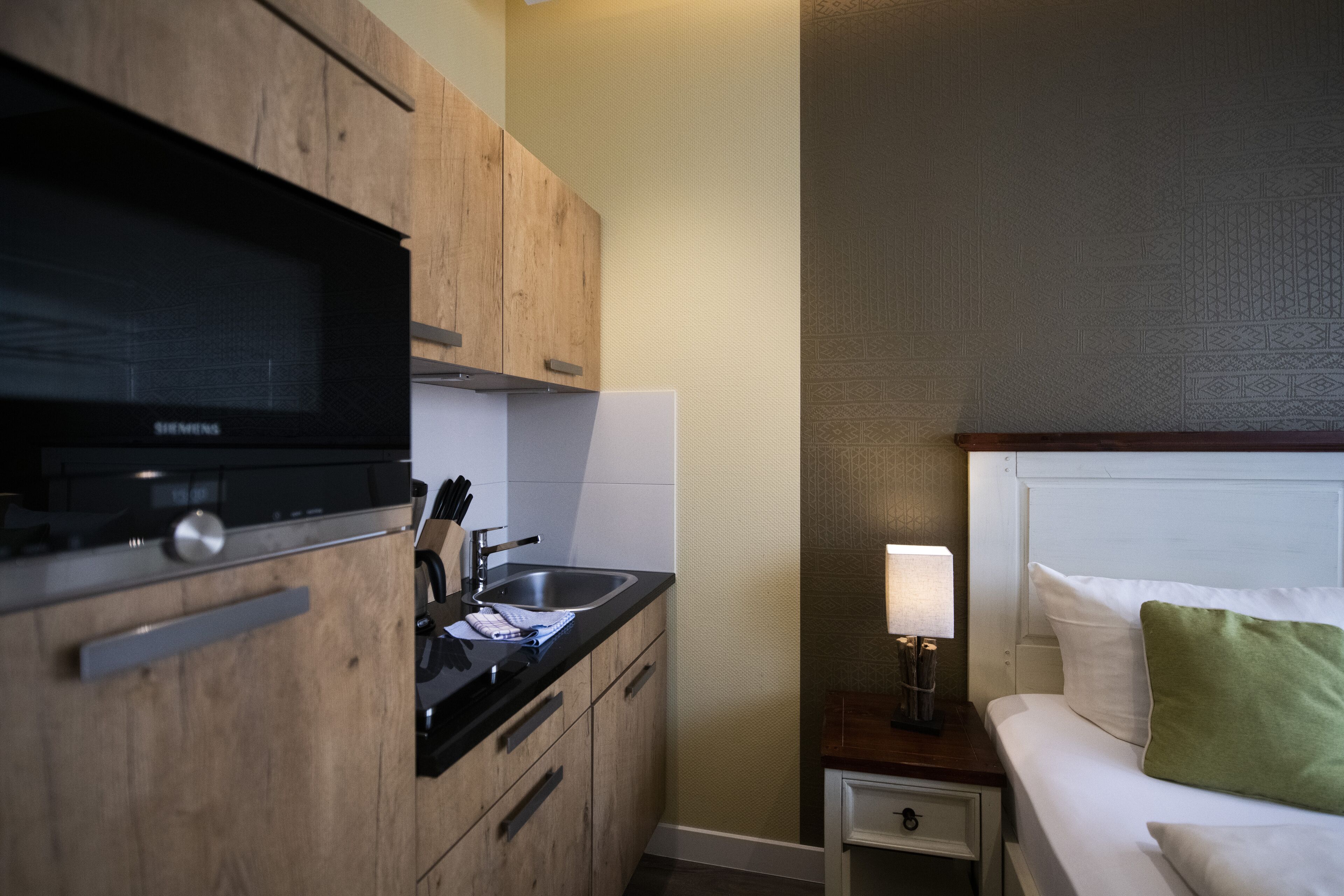 A compact hotel room with a kitchenette, a bed with white bedding and a green pillow, and a bedside lamp.