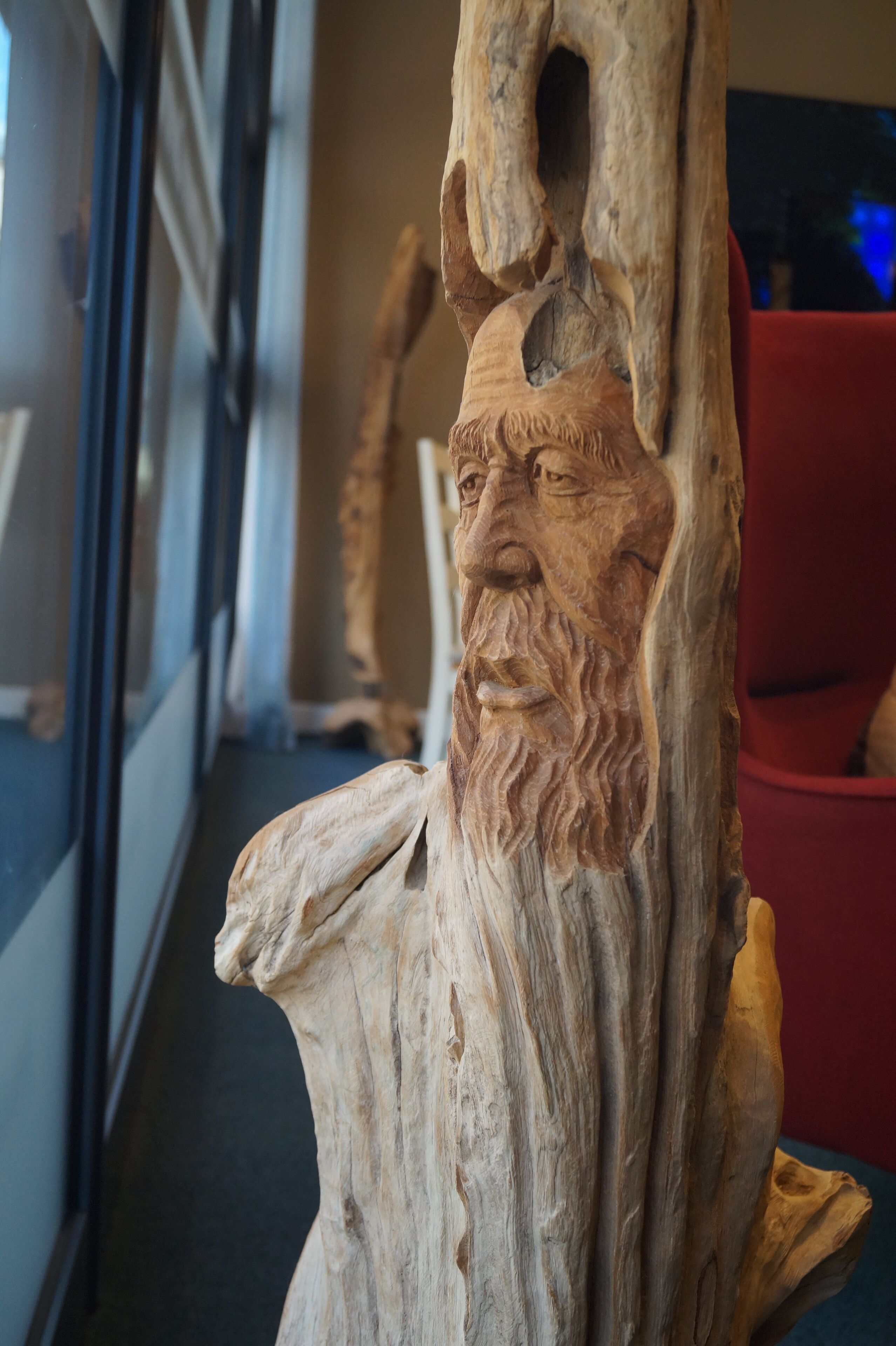 A carved wooden sculpture of a bearded figure with a crown, set against a backdrop of a modern interior with a red chair and a glass door.