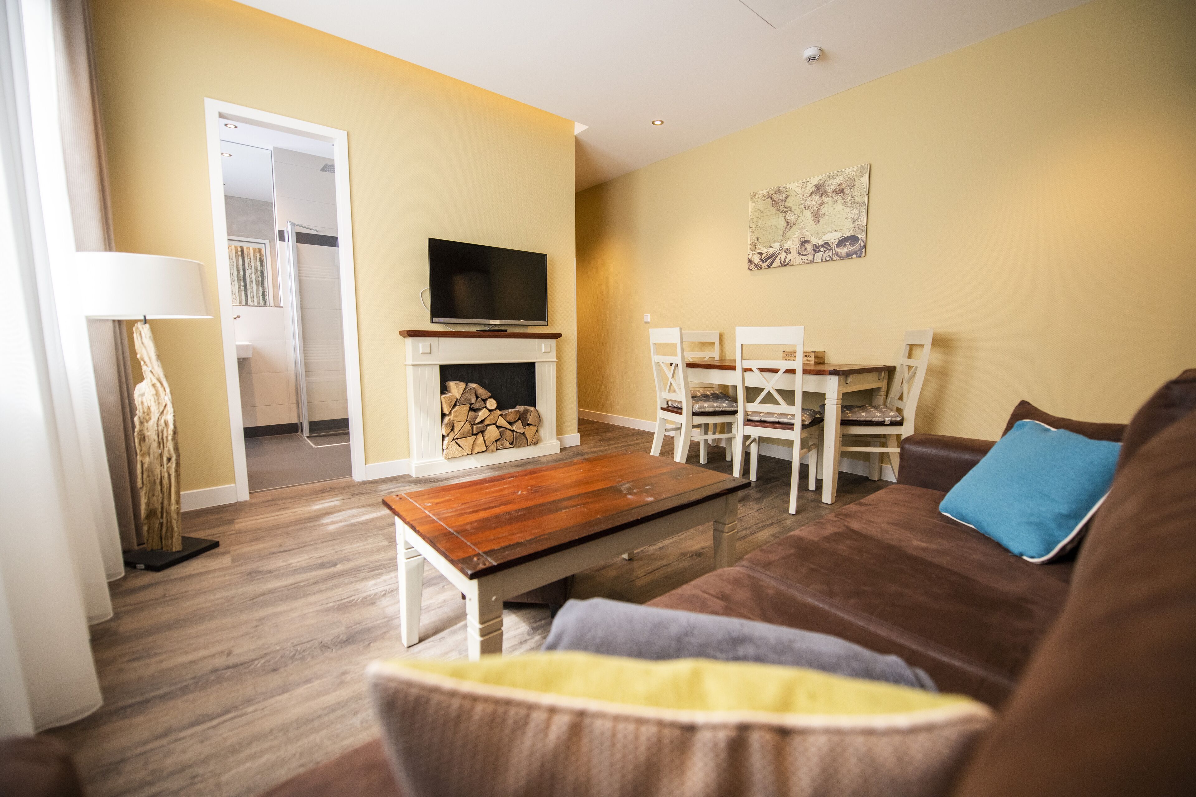 Una sala de estar con un sofá marrón, una mesa de centro de madera y un televisor sobre un mueble. Hay una mesa de comedor con sillas y una chimenea con un televisor encima.