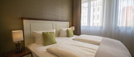 A hotel room with a large bed, two pillows, a nightstand with a lamp, and a window with curtains.