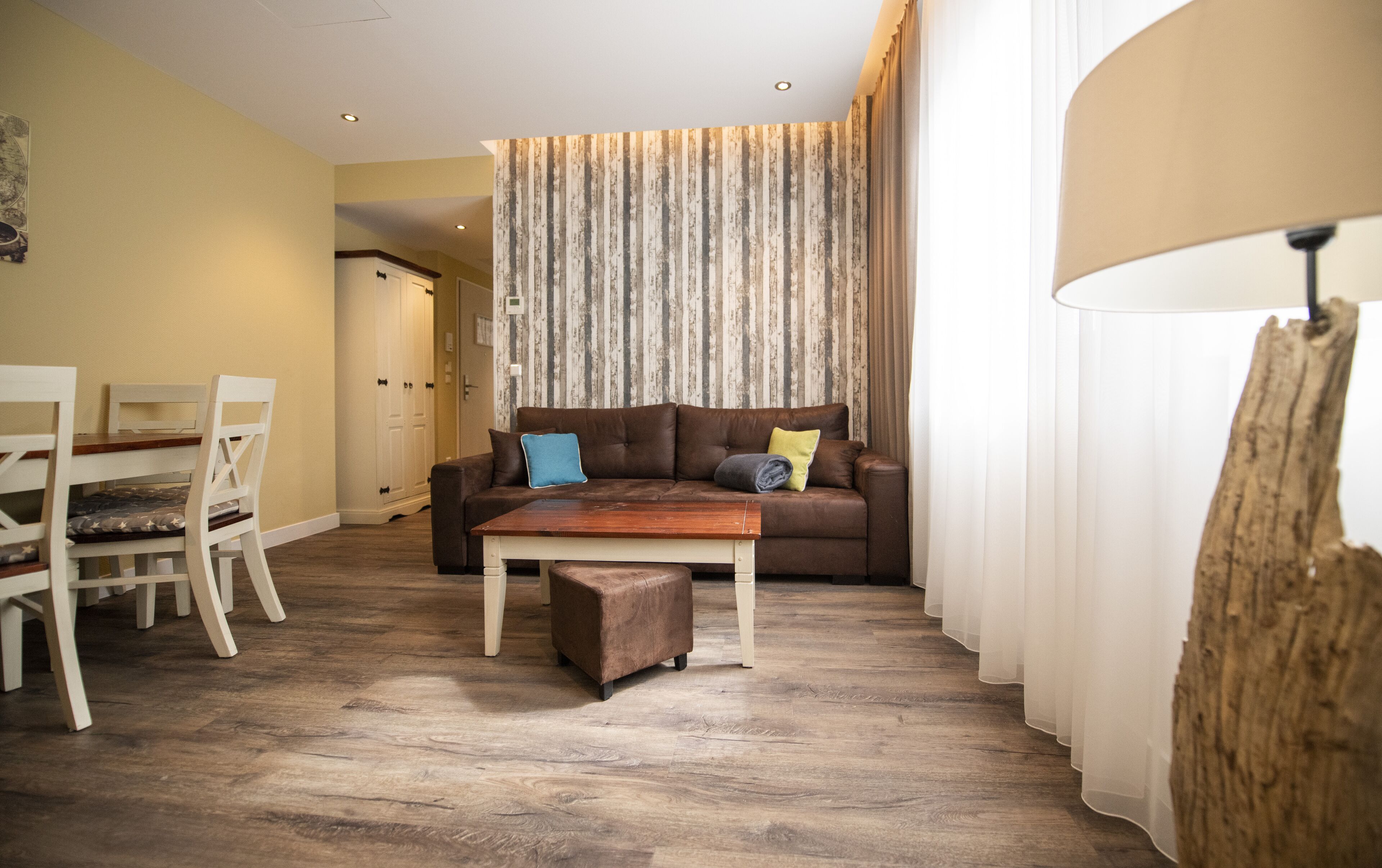 Un salón con un sofá marrón, una mesa de centro de madera y un sillón blanco. Hay una puerta que conduce a otra habitación y una ventana con cortinas traslúcidas.