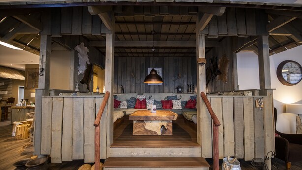 Un interno rustico con una scala in legno che conduce a una zona salotto, travi a vista al soffitto e un orologio appeso al muro.