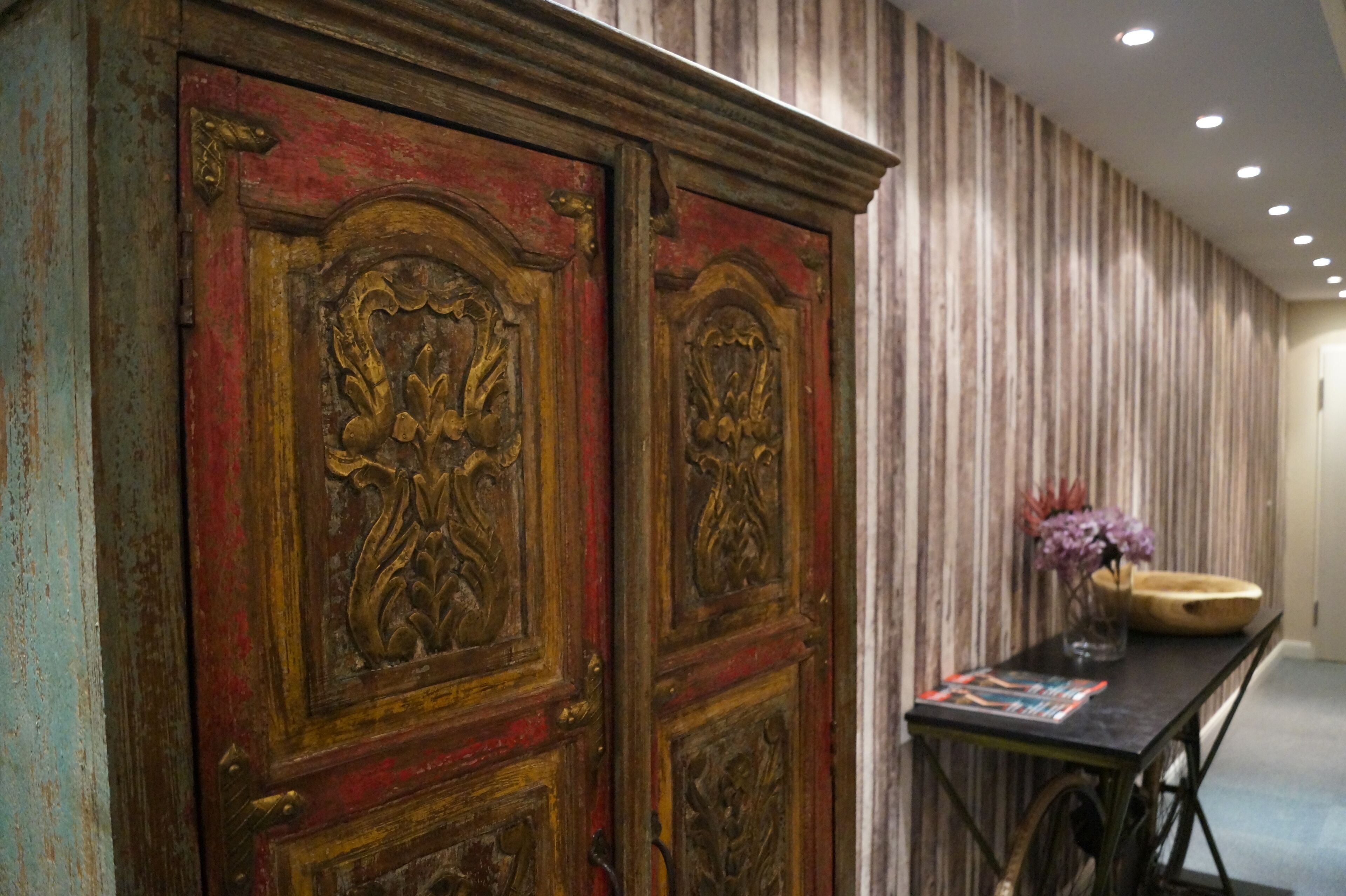 A room with a large wooden cabinet with intricate carvings, a striped wallpaper wall, and a small table with a vase of flowers.