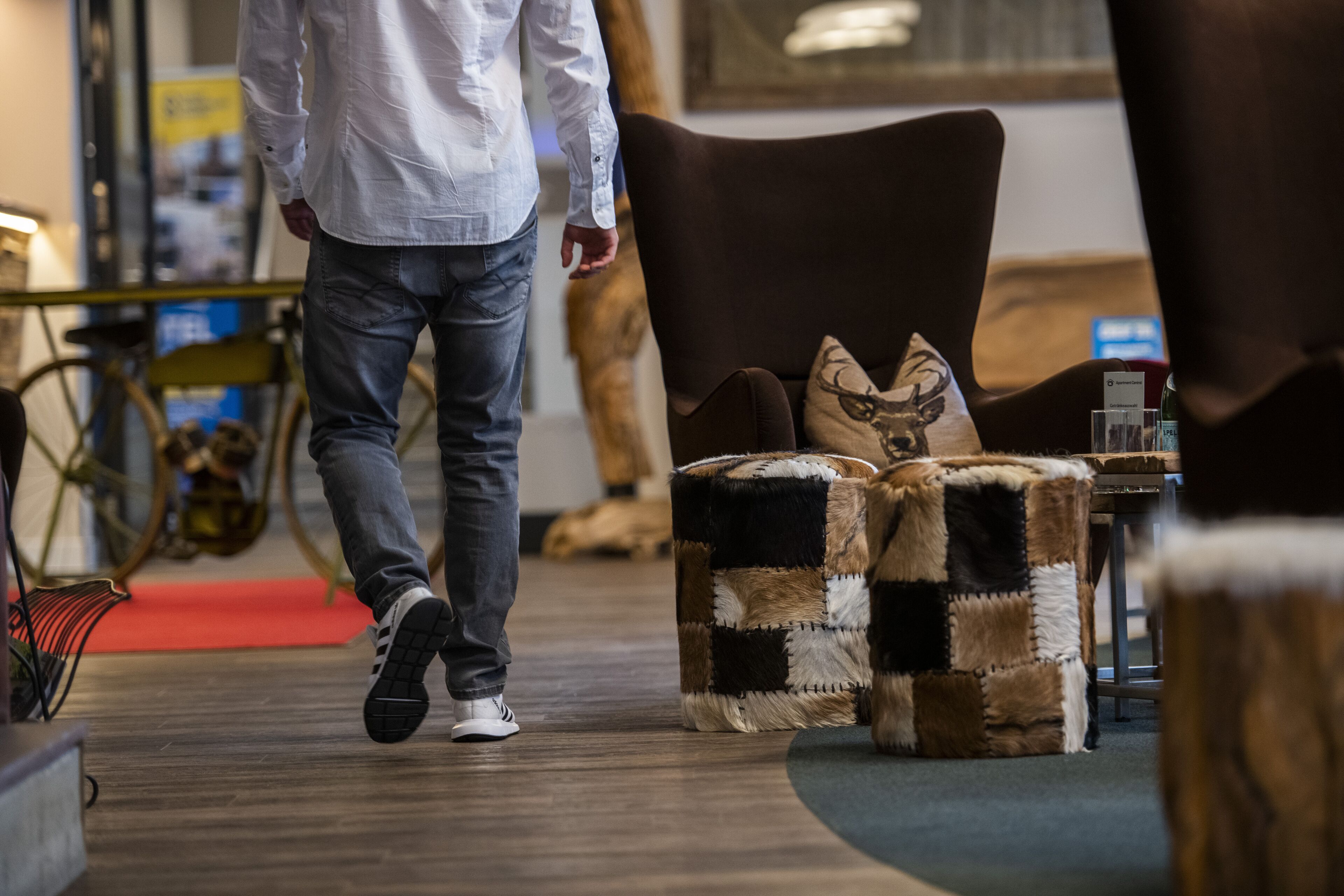 Une personne se promène dans un espace intérieur moderne, doté d’un plancher en bois, d’un fauteuil marron et d’un pouf à carreaux.