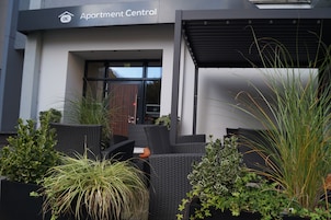 Entrada a un edificio con un letrero que dice 'Apartment Central'. Hay sillas de mimbre y plantas en macetas afuera.