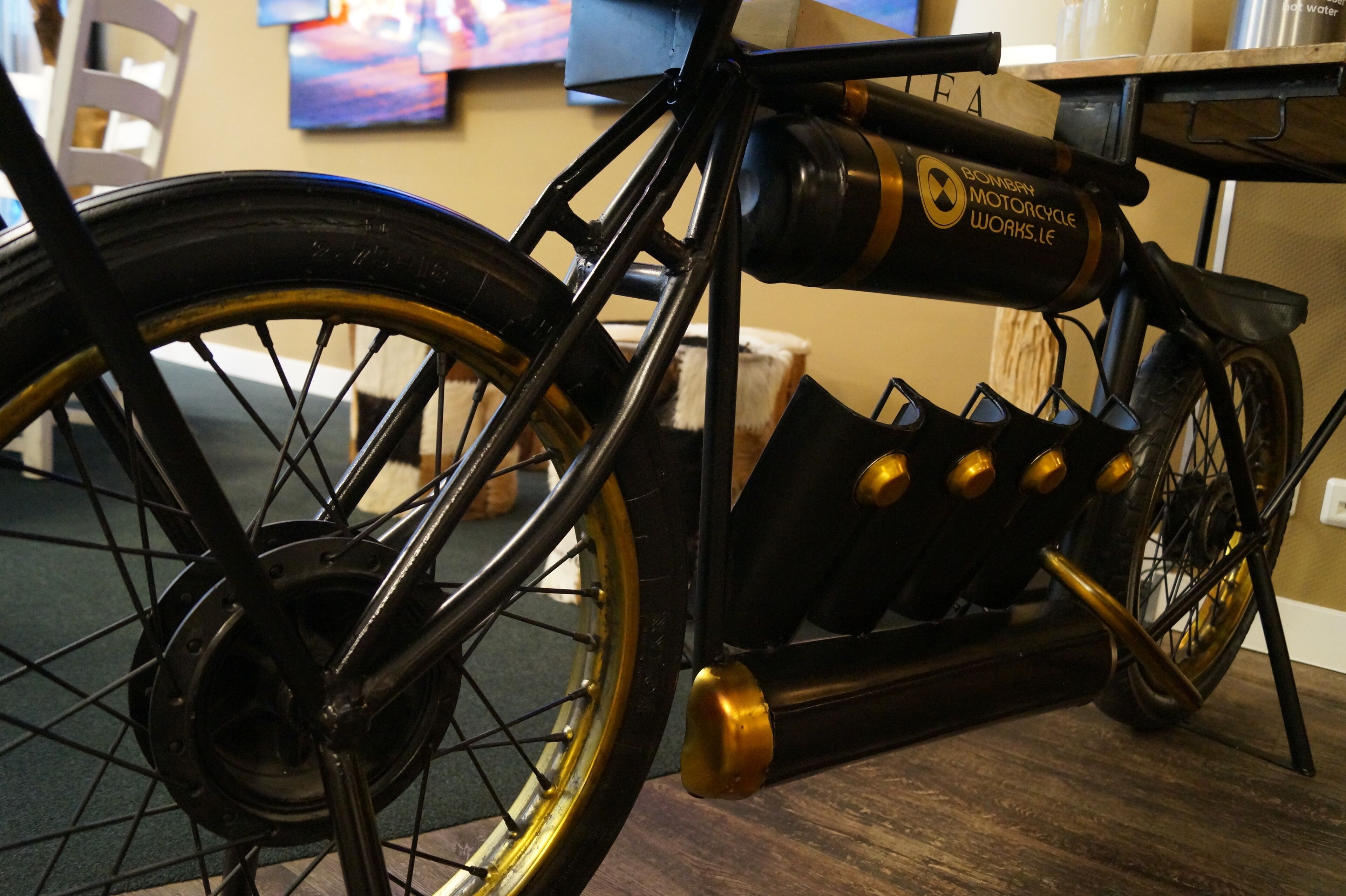 A vintage motorcycle with a black frame and gold accents, displayed indoors with a wooden floor and a painting in the background.