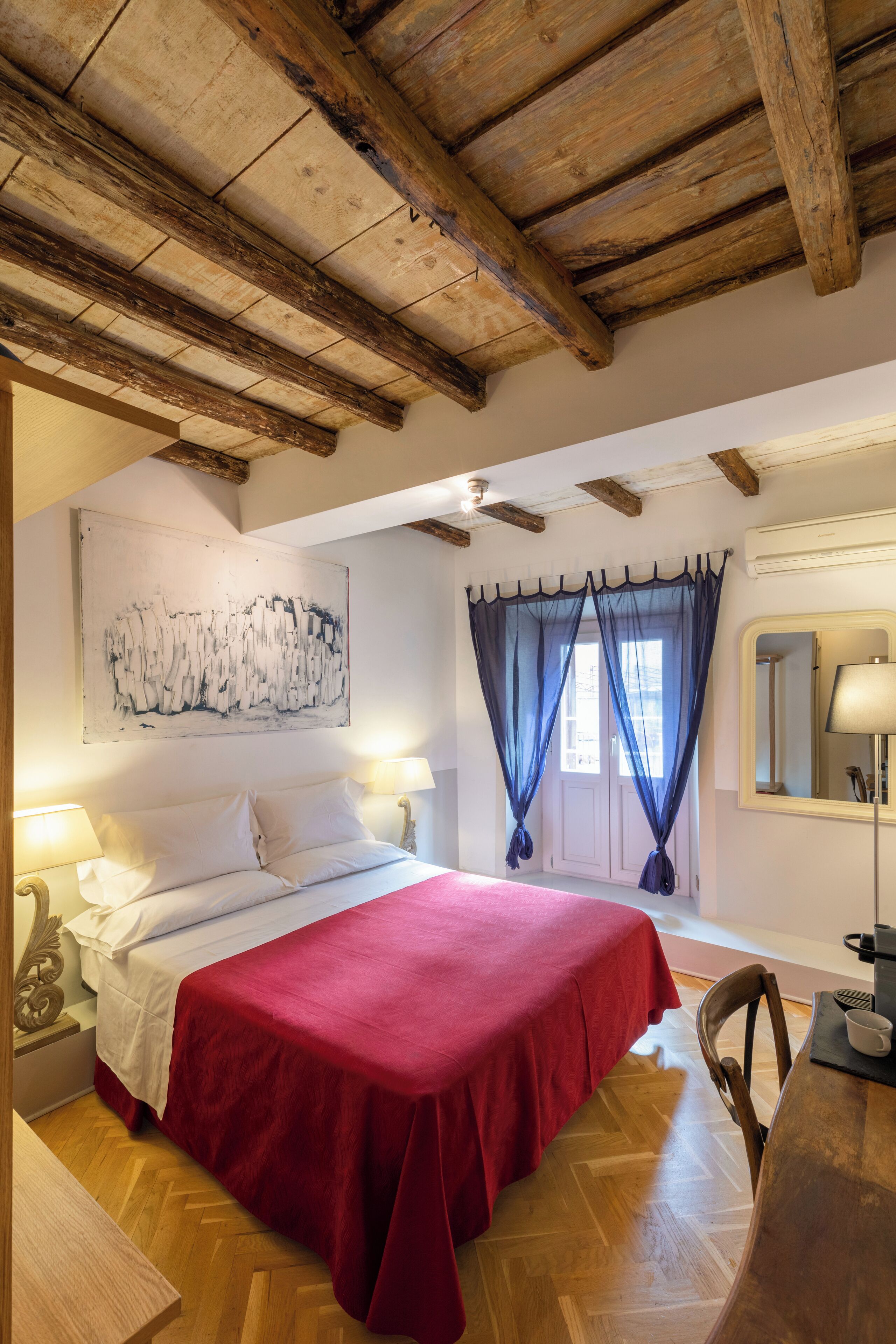 A bedroom with a wooden ceiling, a bed with a red bedspread, a nightstand with a lamp, a chair, and a table with a cup.