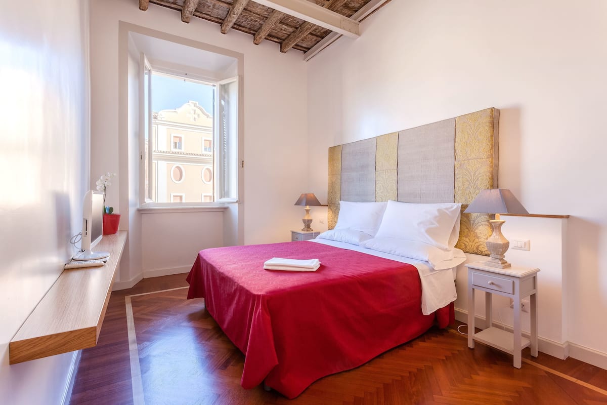 Une chambre avec une couvre-lit rouge, un plancher en bois, une table de chevet avec une lampe et une fenêtre donnant sur des immeubles.