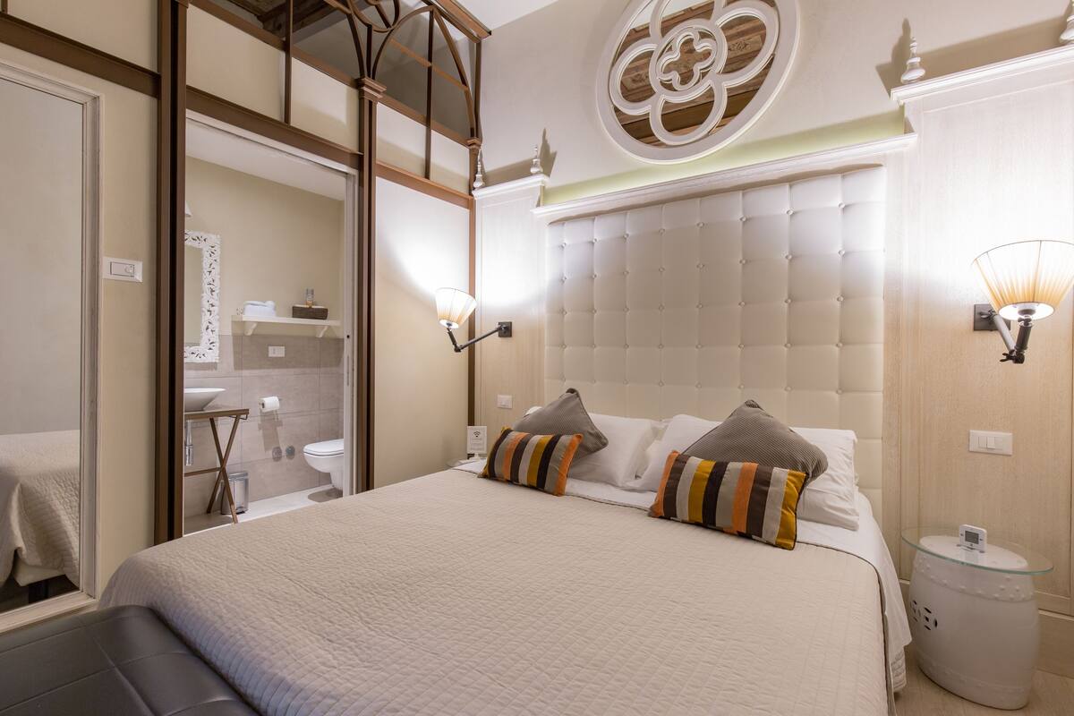 A hotel room with a large bed, a mirror, a bedside table with a lamp, and a wall-mounted light fixture.