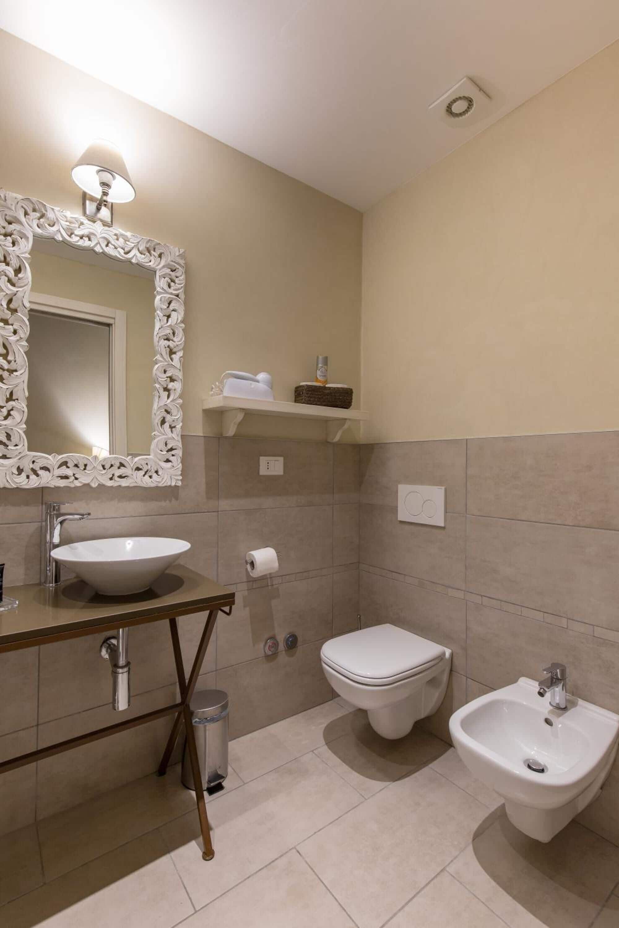 Baño con lavamanos blanco, inodoro y bidé. Se observa un espejo con marco decorativo, un perchero y un estante con artículos de tocador.