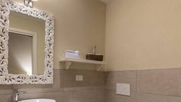 Baño con lavamanos blanco, inodoro y bidé. Se observa un espejo con marco decorativo, un perchero y un estante con artículos de tocador.