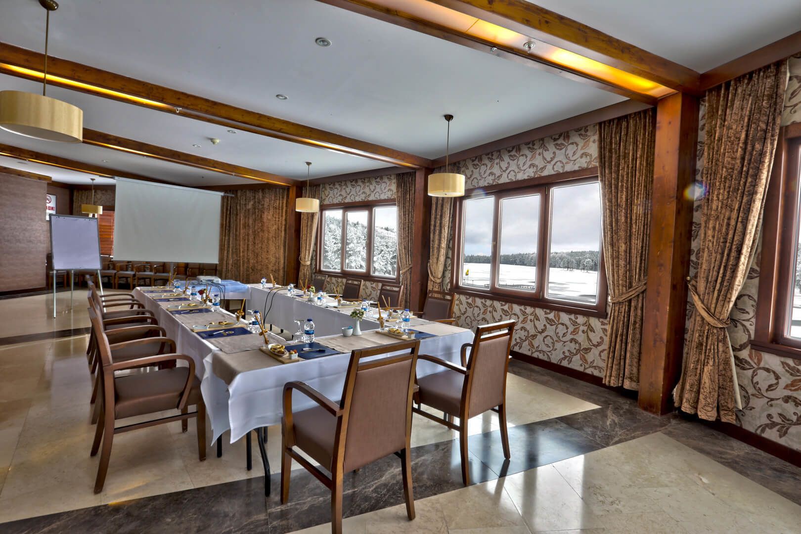 A conference room with a long table, chairs, a projector screen, and large windows with a snowy view.