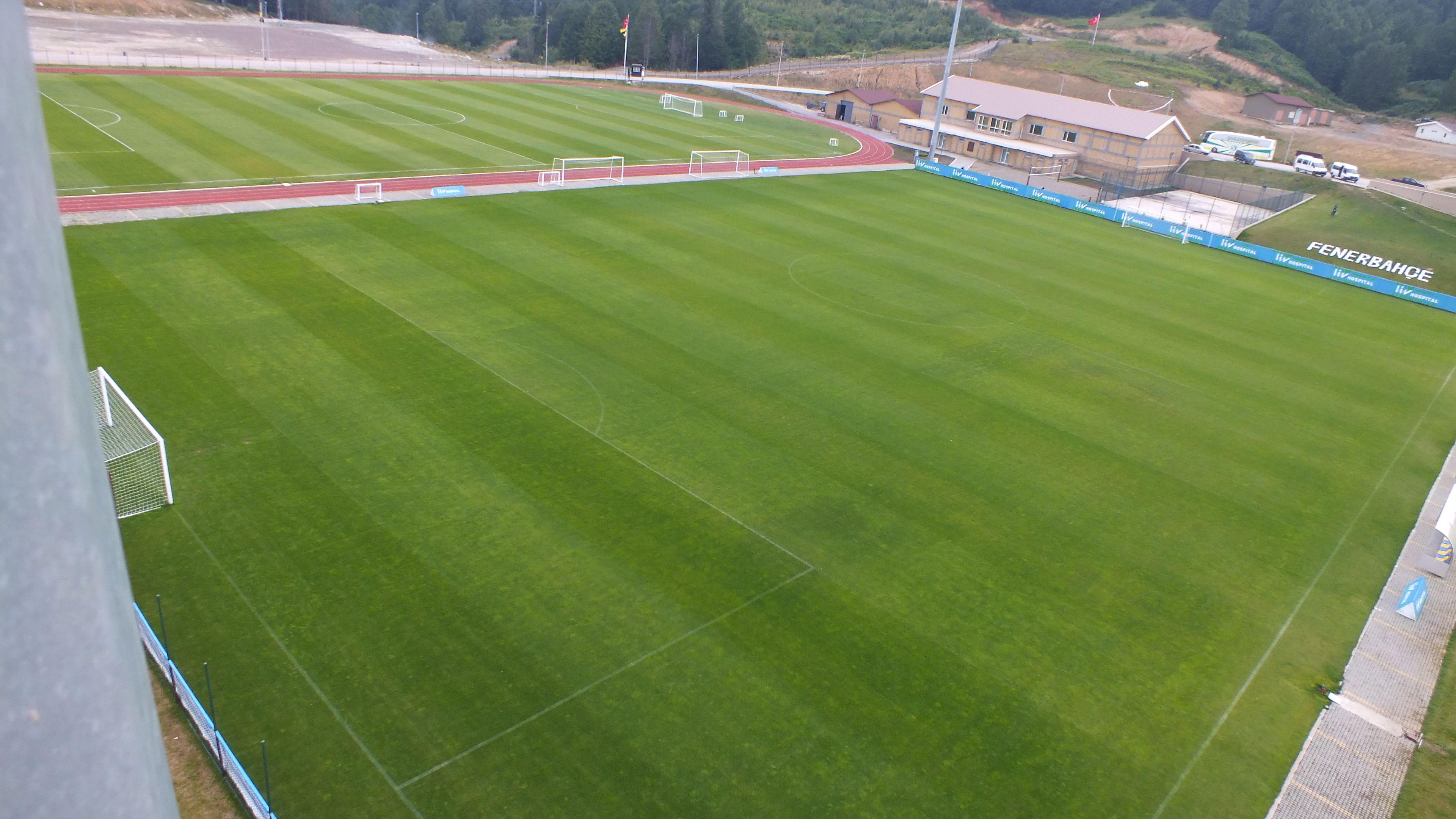 A large, well-maintained sports field with a running track and goalposts, surrounded by a fence and a building with a sign reading "FENERBAHCE".