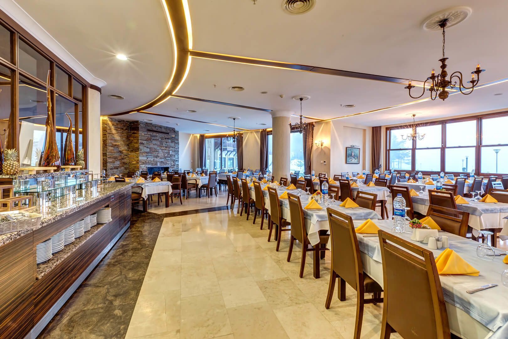 A spacious dining area with multiple tables set for guests, a buffet counter on the left, and large windows providing natural light.