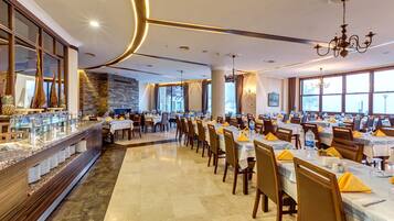 A spacious dining area with multiple tables set for guests, a buffet counter on the left, and large windows providing natural light.
