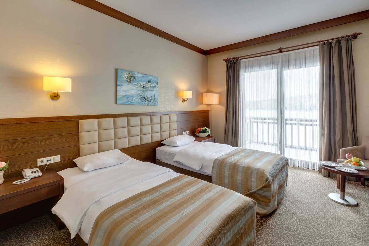 Camera d'albergo con due letti, una testiera in legno, un quadro appeso alla parete, una finestra con tende e un tavolino con una ciotola di frutta.