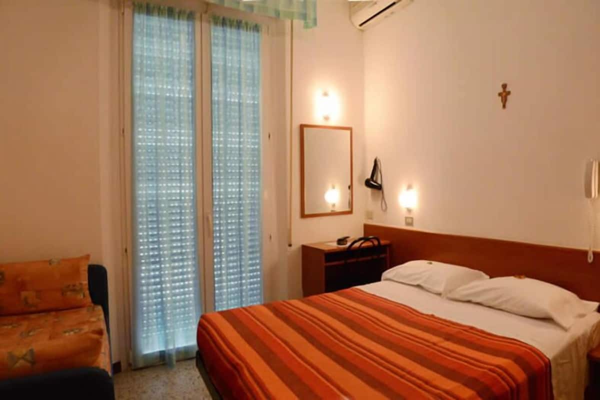 Quarto de hotel com cama, mesa de cabeceira, escrivaninha com abajur, cadeira e janela com persianas.